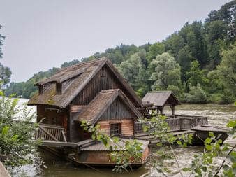 Traditionelle hölzerne schwimmende Mühle mit moosbewachsenen Dächern an der Mur, umgeben von grünen bewaldeten Hügeln.
