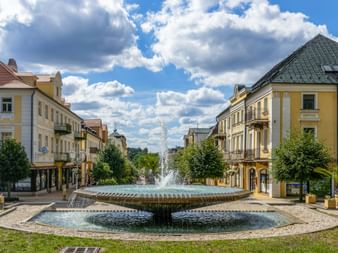 Großer runder Brunnen mit Wasserfontänen an der Národní třída in Franzensbad, umgeben von gelben historischen Gebäuden und Bäumen unter blauem Himmel.