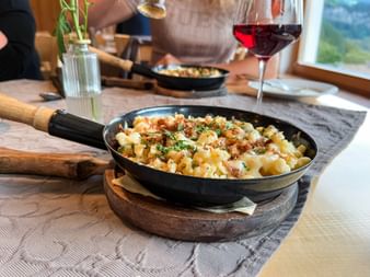 Black pan with Kasnockn pasta topped with herbs and breadcrumbs on wooden board. Red wine glass and second pan visible in background.