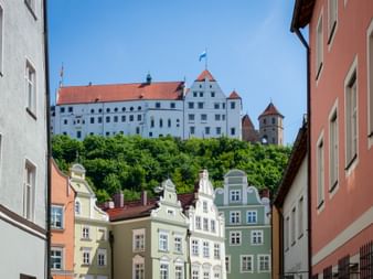 Blick durch bunte historische Häuser in Landshut zur Burg Trausnitz auf einem Hügel. Das weiße Schloss mit roten Dächern erhebt sich über grünen Bäumen.