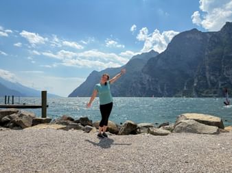 Woman in light blue shirt with outstretched arms on rocky shore at Lake Garda near Riva, with mountains and blue sky in background.