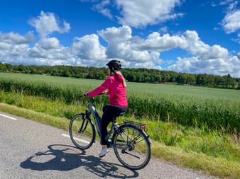 Frau in rosa Jacke radelt auf asphaltierter Straße durch grüne Felder unter blauem Himmel mit weißen Wolken bei Hamburg-Lübeck.