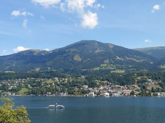 Passenger ferry on Millstätter See with lakeside town and green mountains under blue sky. Alpine landscape along Alpe-Adria cycle route.