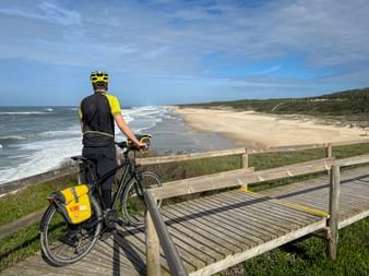 Cyclist with yellow panniers on wooden boardwalk overlooking sandy beach and Atlantic Ocean on Portugal's Alto Minho coastal path.