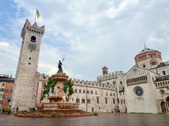 Piazza Duomo in Trient with Neptune Fountain, medieval clock tower, and cathedral. Historic buildings surround the cobblestone square under a cloudy sky.