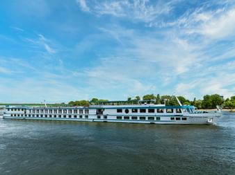 White river cruise ship MS Switzerland sailing on calm water with blue sky. The multi-deck vessel features rows of windows and a sun deck.