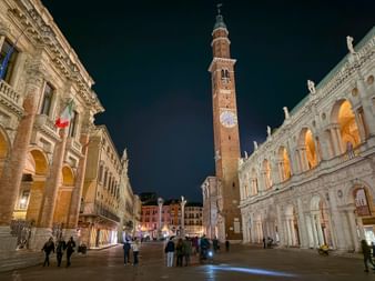 Night view of Piazza dei Signori in Vicenza with illuminated brick bell tower, Renaissance buildings, and Italian flag on the left facade.