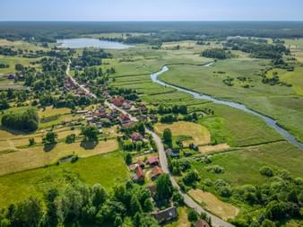 Luftaufnahme eines kleinen Dorfes in Masuren mit roten Dächern, grünen Wiesen, einem gewundenen Fluss und einem See in der Ferne.
