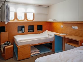 Modern ship cabin on MS Ave Maria with two single beds, wooden paneling, three porthole windows, storage compartments, and white bedding.