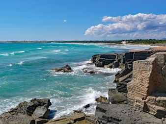 Felsige Küste am Strand Es Trenc auf Mallorca mit türkisfarbenem Meer, Wellen an geschichteten Felsen, Sandstrand und Vegetation im Hintergrund.