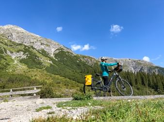 Female cyclist with yellow panniers on e-bike pausing on Formarinweg near Dalaas, with green alpine slopes and rocky mountain peaks under blue sky.