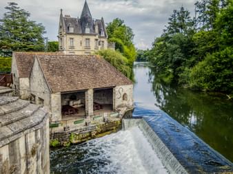 Old stone mill with water weir on a calm river in Moret-sur-Loing, with a château featuring a pointed tower visible behind, surrounded by lush trees.