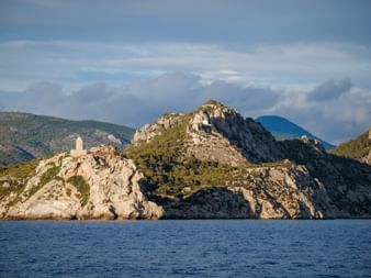 Weißer Leuchtturm auf felsiger Halbinsel im Golf von Gökova mit Bergen im Hintergrund. Blaues Meer im Vordergrund unter teilweise bewölktem Himmel.