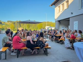 Mitarbeiter beim Eurofun Touristik Sommerfest sitzen an Holzbiergarnituren im Innenhof, mit Gebäude und grünen Hügeln im Hintergrund.