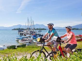 Zwei Radfahrer fahren am Chiemsee in Gstadt entlang, Boote liegen im Hafen, Berge im Hintergrund unter blauem Himmel.