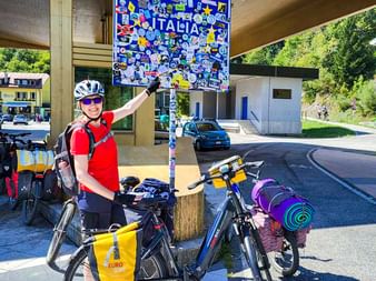 Radfahrerin in rotem Trikot zeigt auf blaues Italia-Grenzschild mit Aufklebern. Zwei Tourenräder mit gelben Taschen stehen daneben.
