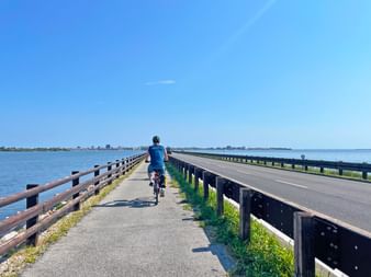 Radfahrer auf separatem Radweg entlang Brückendamm in Grado, Friaul. Blaue Lagunenwasser auf beiden Seiten, klarer Himmel darüber.