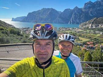 Two cyclists in helmets taking a selfie with Lake Garda, Torbole town, and dramatic mountains in the background under blue sky.