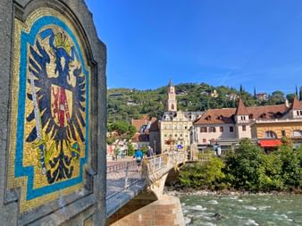 Steinbrücke in Meran mit Mosaik-Reichsadler-Wappen. Historische Stadtgebäude und Kirchturm über dem Fluss unter blauem Himmel sichtbar.