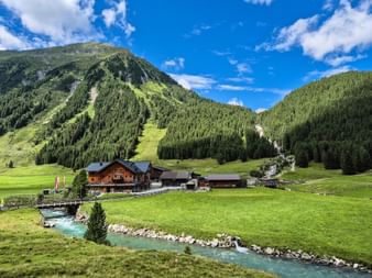 Traditional wooden alpine lodge in Pinzgau valley with turquoise mountain stream, green meadows, and forested peaks under blue sky.