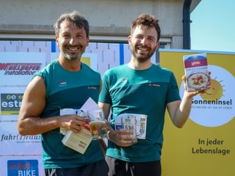 Two smiling men in turquoise Vaude sports shirts holding food products at Grabenseelauf 2026, standing in front of sponsor banners.