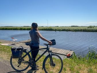 Female cyclist with touring bike standing by a wooden dock on a canal. Green fields and reeds visible across the water under blue sky.