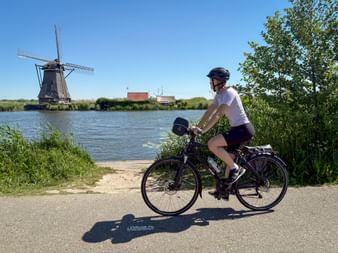 Radfahrerin auf einem Weg neben einem Kanal mit traditioneller holländischer Windmühle in Kinderdijk unter blauem Himmel.