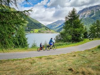 Erwachsener und Kind radeln auf Weg am Reschensee mit Blick auf Graun, Bergen und Nadelbäumen unter bewölktem Himmel.