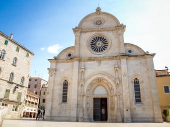 Facade of the Cathedral of St. James in Šibenik, Dalmatia, with ornate rose window and arched entrance under blue sky.
