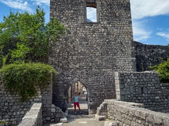 Steinerner Torturm Porta San Genesio in Venzone mit Radfahrer im Torbogen. Mittelalterliche Mauern und grüne Bäume unter blauem Himmel.