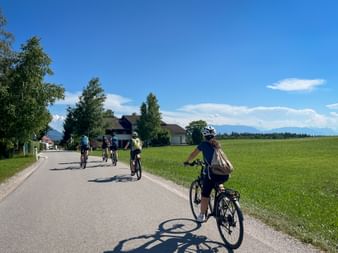 Group of cyclists on a rural road with green fields, houses, and mountains in the background under a blue sky with white clouds.