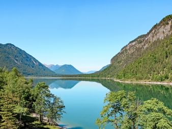 Sylvenstein Reservoir surrounded by forested mountains under clear blue sky. The turquoise water reflects the mountains and trees perfectly.