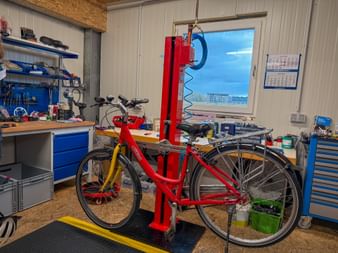 Rotes Fahrrad auf Reparaturständer in Werkstatt mit blauen Werkzeugschränken, Werkbank und Fenster mit Außenblick in Schärding.