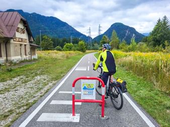 Radfahrer in gelber Jacke am Alpe Adria Radweg mit Ciclovia-Schild, Berge und altes Gebäude sichtbar. Region Kanaltal.