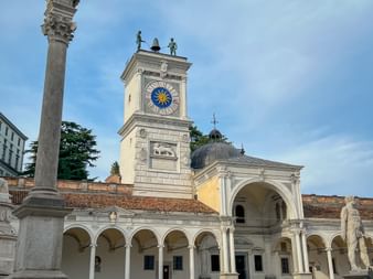 Historischer Uhrturm mit blau-goldenem Zifferblatt an der Piazza della Libertà in Udine mit weißer Steinarchitektur, Arkaden und Statuen.