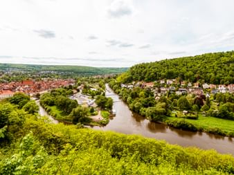 Aerial view of Hann. Münden showing rivers flowing through green landscape with red-roofed town and forested hills under cloudy sky.