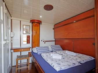 Gemütliche Kabine auf der MS Macek mit französischem Bett, blau-weiß gestreiften Kissen, Holzverkleidung, Schminktisch und maritimer Dekoration.