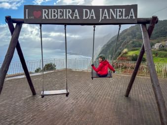 Cyclist in red jacket on swing under sign reading 'I LOVE RIBEIRA DA JANELA' with ocean and green hills in background on Madeira.