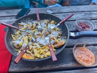 Large pan of torn Kaiserschmarrn pancake dusted with powdered sugar, surrounded by forks with red handles and two bowls of jam on wooden table.
