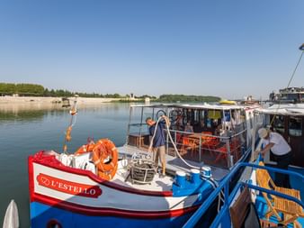 MS L'Estello Boot mit blau-weißem Rumpf am ruhigen Fluss vertäut. Besatzungsmitglieder an Deck, oranger Rettungsring sichtbar. Sandiges Ufer und Bäume im Hintergrund.
