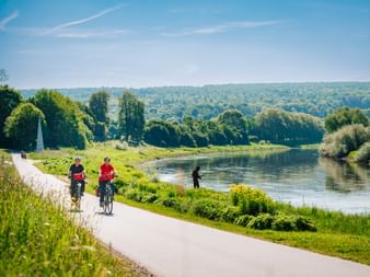 Zwei Radfahrer in roten Shirts fahren auf einem asphaltierten Weg entlang der Weser. Grüne Wiesen und Bäume säumen das Flussufer unter blauem Himmel.