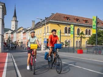 Two cyclists with panniers riding on a street in Villach. Historic buildings and a church tower are visible under a blue sky.