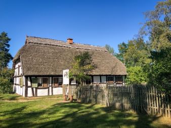 Half-timbered house with thick thatched roof and wooden fence in Kluki. White walls with dark beams, surrounded by green lawn and trees under blue sky.