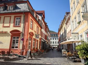Kopfsteinpflasterstraße in Heidelbergs Altstadt mit bunten historischen Gebäuden. Außengastronomie auf beiden Seiten unter blauem Himmel.