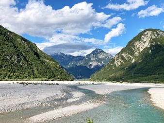 Türkisfarbener Fluss mit weißen Kiesbänken fließt durch ein Bergtal. Grüne bewaldete Hänge erheben sich beiderseits unter blauem Himmel mit Wolken.