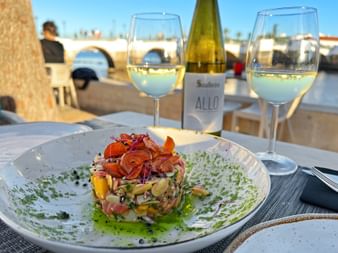 Bunter Fischtartar mit knusprigem Schinken auf weißem Teller, serviert mit zwei Gläsern Weißwein und einer Flasche 'Allo' im Minho.