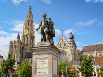 Bronzestatue von Peter Paul Rubens auf Steinsockel mit gotischem Turm der Liebfrauenkathedrale im Hintergrund unter blauem Himmel in Antwerpen.