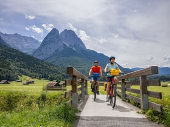 Two cyclists crossing a wooden bridge on a bike path near Garmisch with dramatic mountain peaks and green meadows in the background.