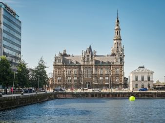 Historisches Lotsenhaus mit verziertem Turm in Antwerpen, über einen Kanal mit gelber Boje hinweg betrachtet. Moderne und traditionelle Gebäude säumen das Ufer.