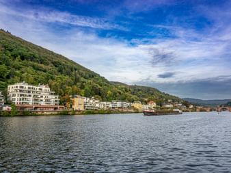 Blick auf Heidelberg am Neckar mit modernen und historischen Gebäuden vor bewaldeten Hügeln unter blauem bewölktem Himmel.
